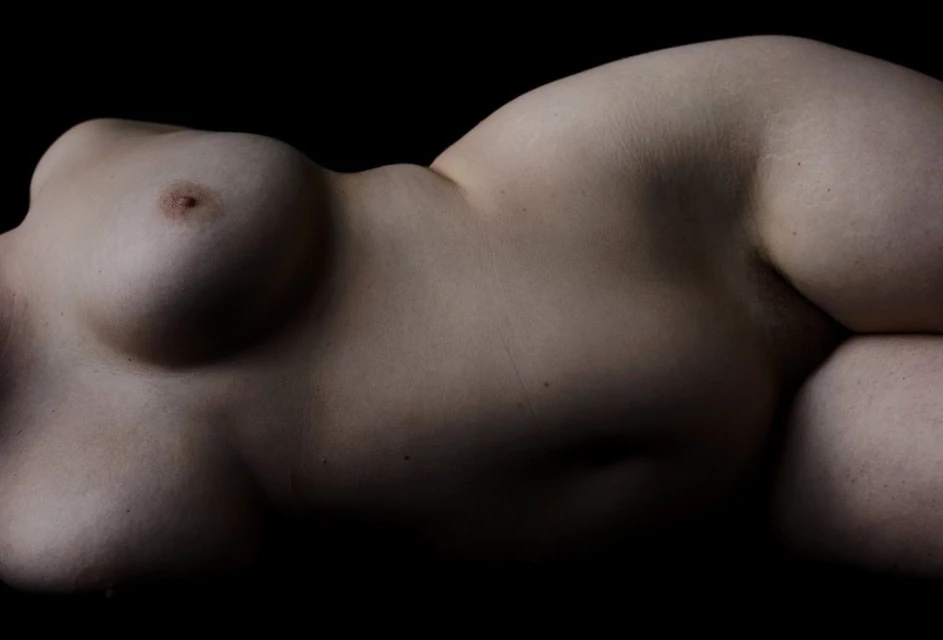 Nude torso of an adult female lying on her side in a dimly lit black studio with a full view of her breast and midsection.