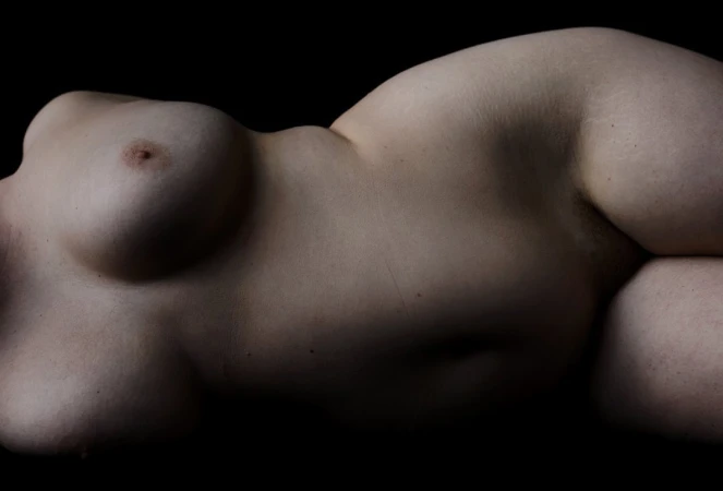 Nude torso of an adult female lying on her side in a dimly lit black studio with a full view of her breast and midsection.