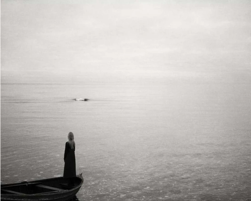 A solitary woman with long blonde hair stands in a small boat on a misty sea, gazing out at the horizon on a cloudy day in black and white.