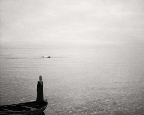 A solitary woman with long blonde hair stands in a small boat on a misty sea, gazing out at the horizon on a cloudy day in black and white.