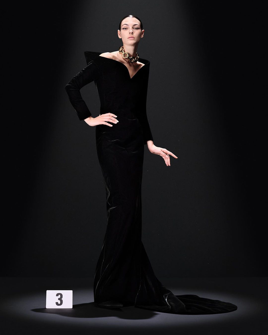 Elegant woman in a long black gown poses in a spotlight with a gold necklace, fashion forward, artistic studio shot.