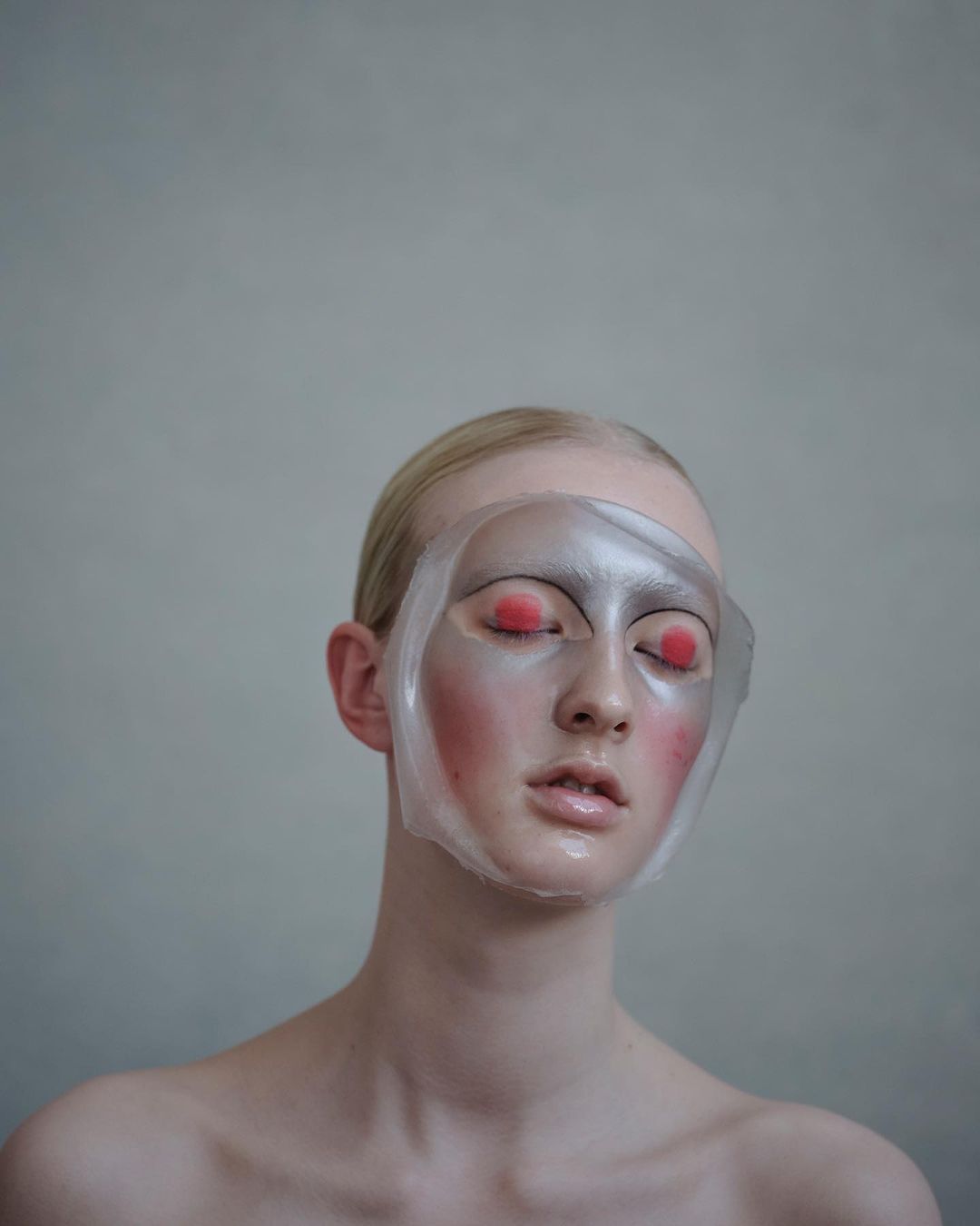 Close-up of a fair-skinned person with a clear face mask, red eye makeup, and neutral expression against a light gray background.