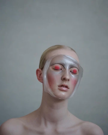 Close-up of a fair-skinned person with a clear face mask, red eye makeup, and neutral expression against a light gray background.