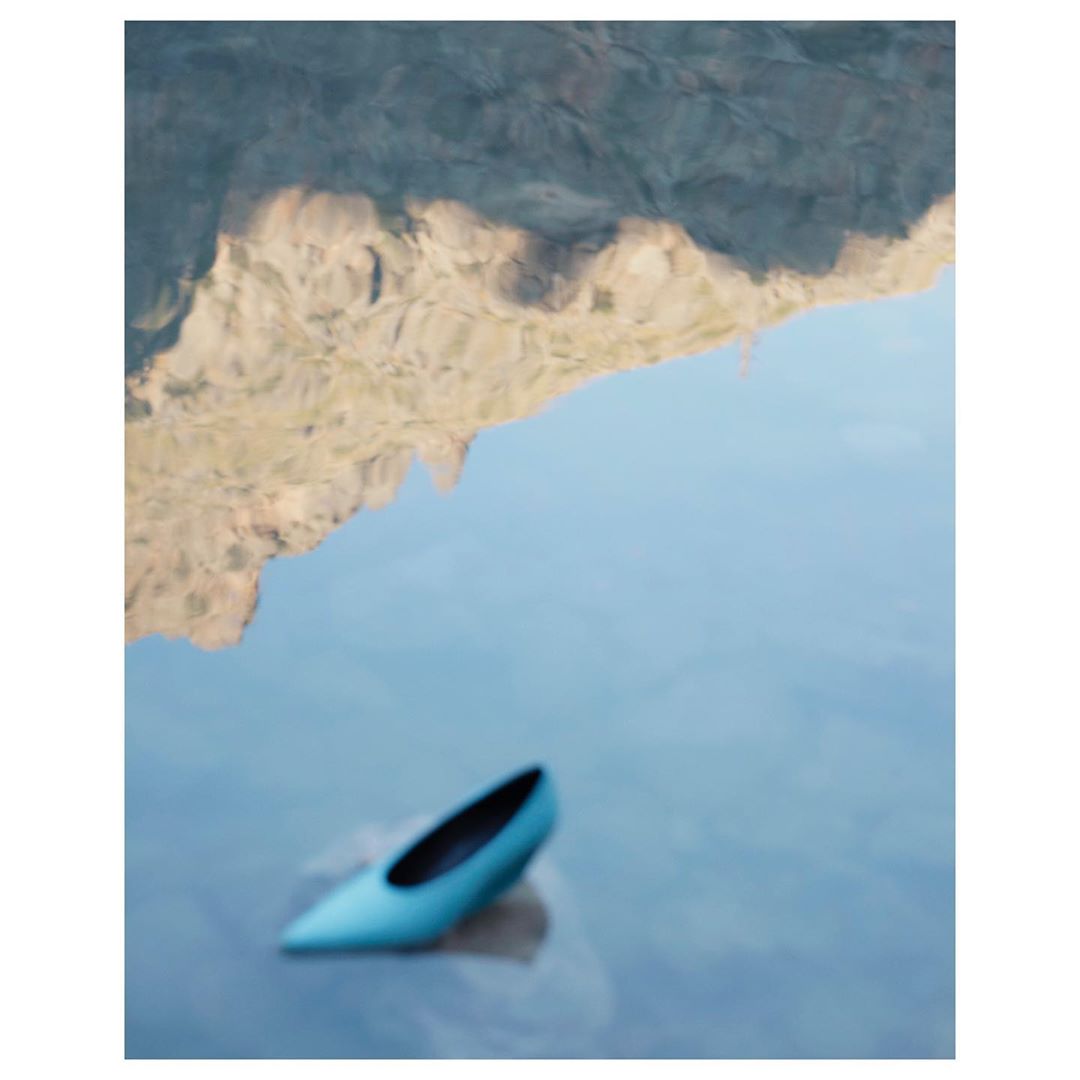 Teal high heel sits on water surface, reflected mountains visible. Conceptual image, possibly representing fashion and the natural world.