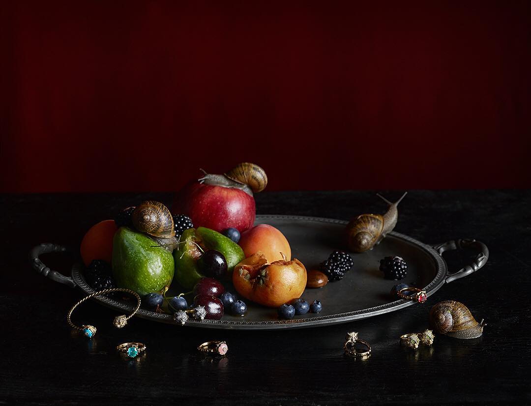Still life of fruit on a silver tray, decorated with snails and rings, set against a dark red background, in dim studio lighting.