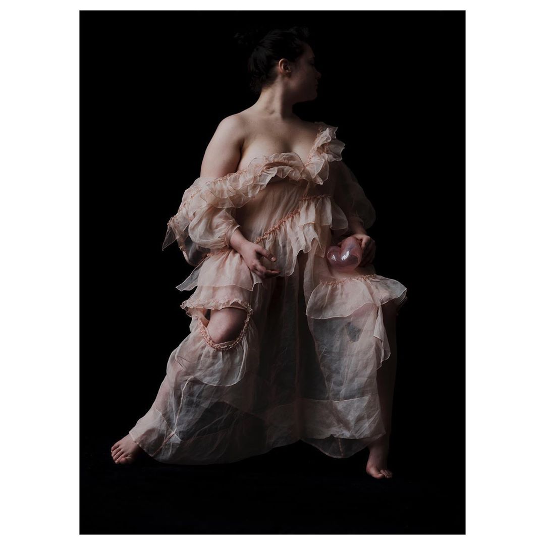Woman poses in a ruffled dress, seated, and holding a glass orb against a black background in a studio photography setting.