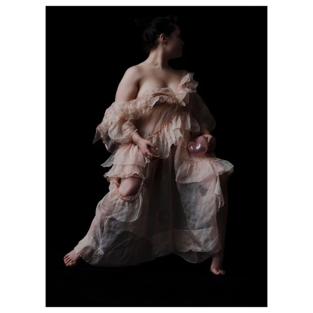Woman poses in a ruffled dress, seated, and holding a glass orb against a black background in a studio photography setting.