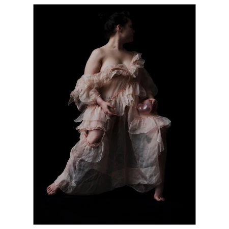Woman poses in a ruffled dress, seated, and holding a glass orb against a black background in a studio photography setting.
