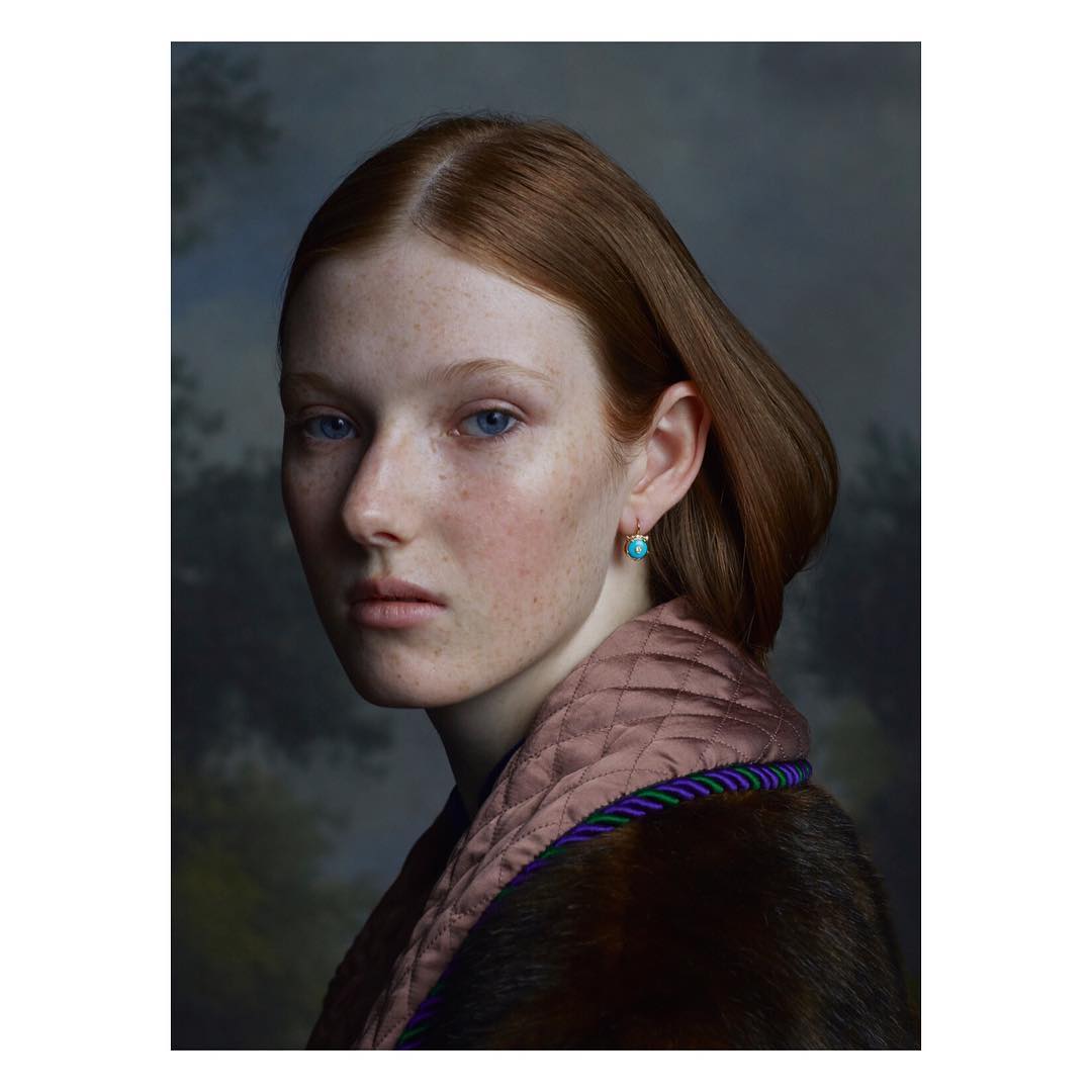 Portrait of a red-haired woman with fair skin and freckles, wearing a fur-trimmed robe, against a blurred, painterly background.