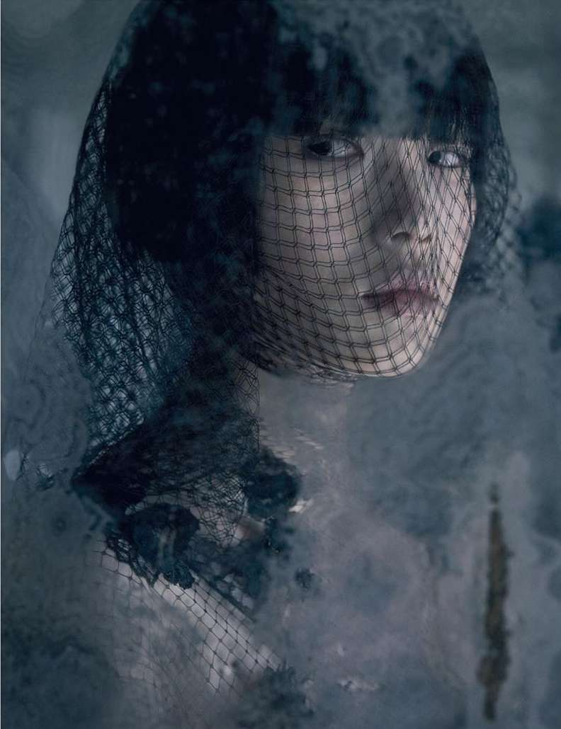Portrait of a woman with dark hair and a net veil over her face, set against a hazy, muted blue-gray backdrop in soft studio light.