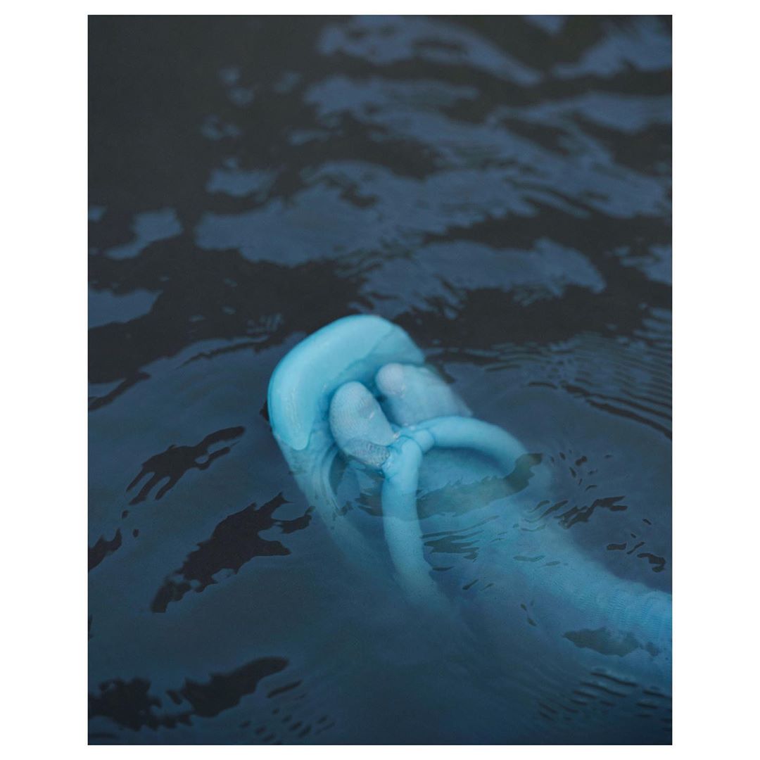 A blue foot and toes float in dark blue water, creating ripples, suggesting an underwater perspective with cool tones and surreal atmosphere.