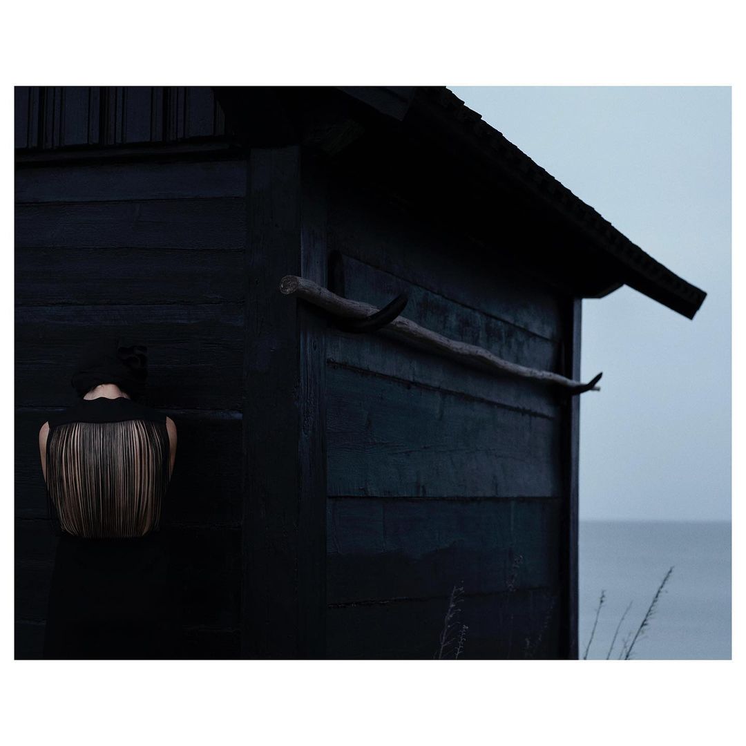 Artful image of a woman in a textured top standing next to a dark wooden structure, evoking a sense of mystery and connection with nature.