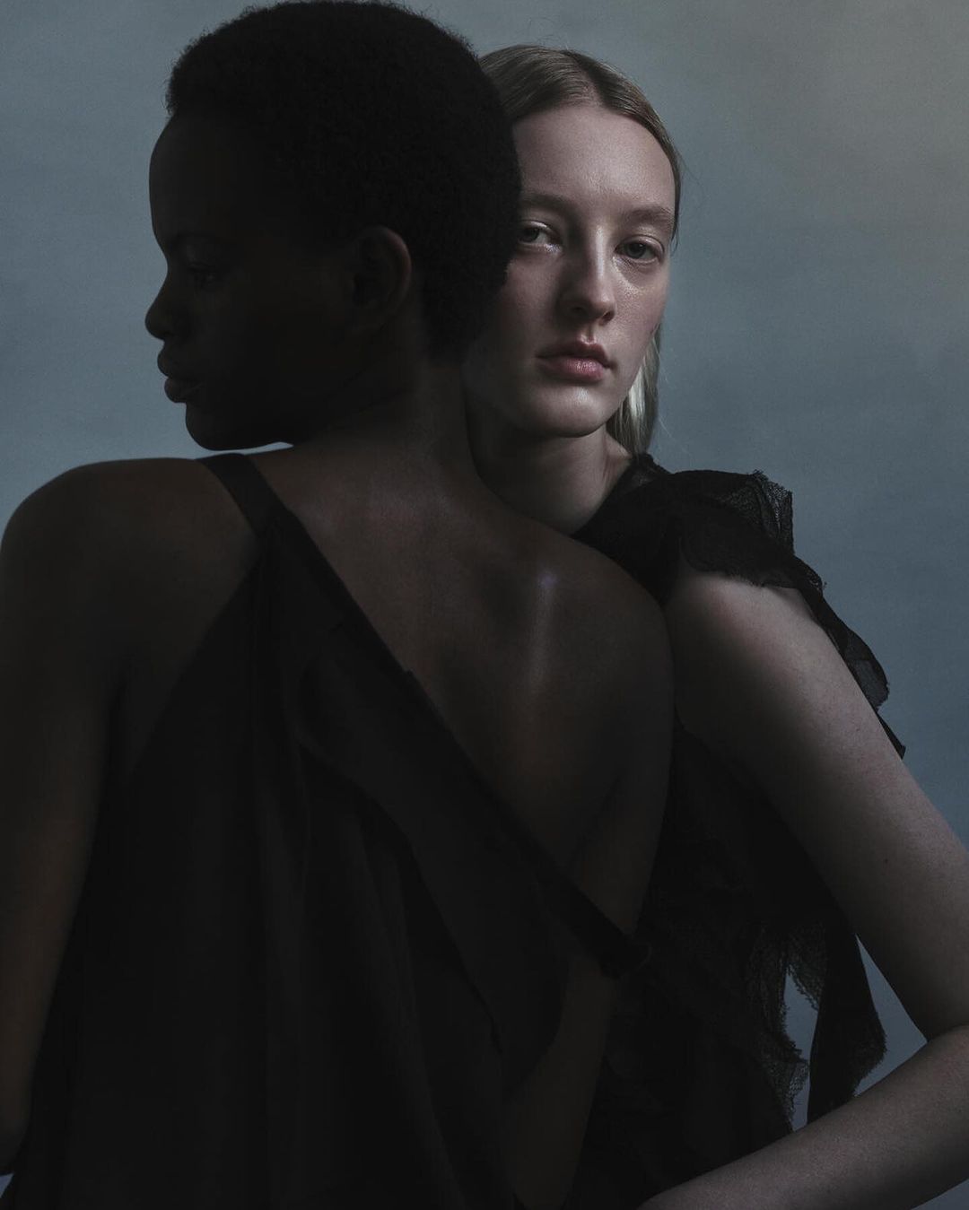 Two women posing closely together in moody lighting, one with dark skin and one with fair skin wearing dark dresses.