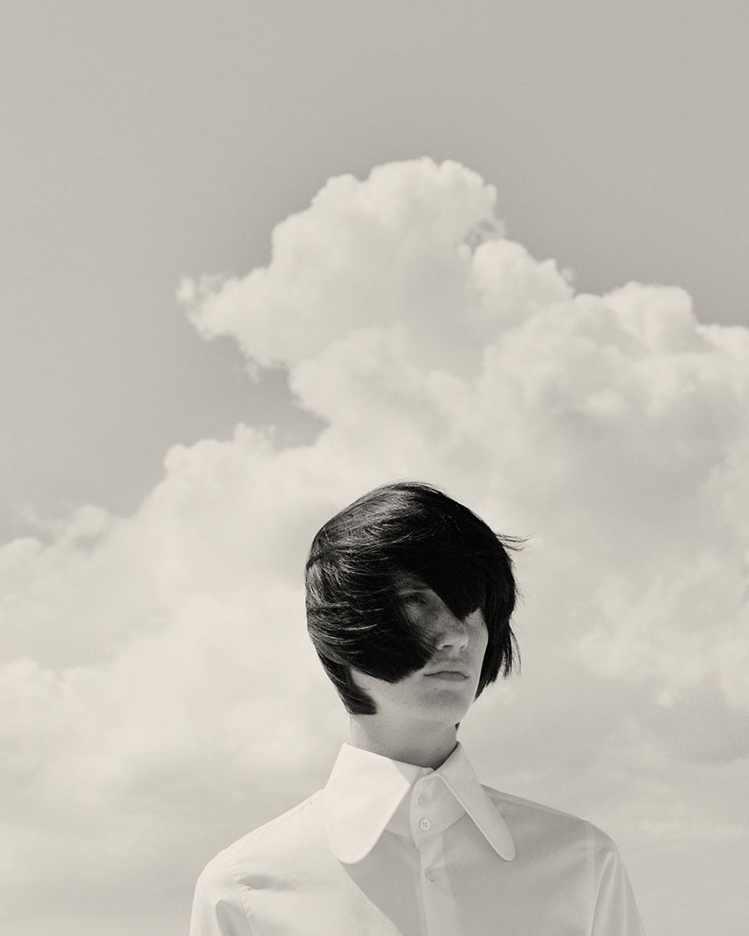 Black and white portrait of a person with dark hair and a white collared shirt against a backdrop of fluffy clouds.