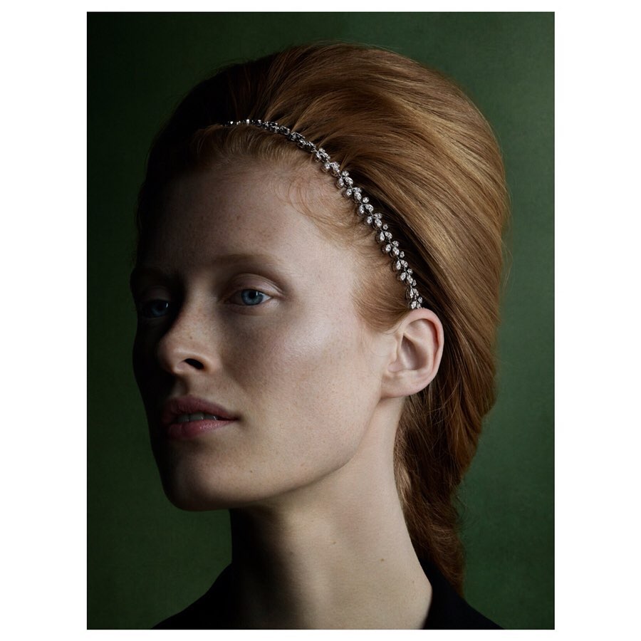Close-up portrait of a red-haired woman with fair skin wearing a decorative headband against a dark green background.