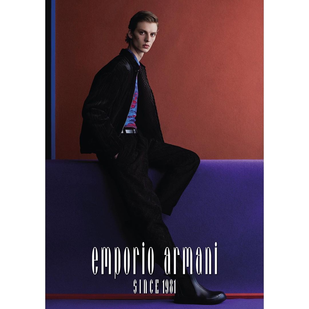 A male model posing against a purple ledge in front of an orange background wearing a black jacket and patterned shirt.
