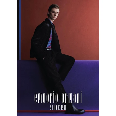 A male model posing against a purple ledge in front of an orange background wearing a black jacket and patterned shirt.
