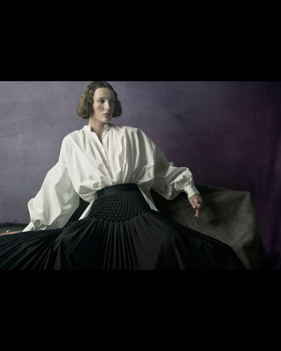 A poised female fashion model in a white blouse and a black skirt poses in an indoor setting with a dark purple backdrop.