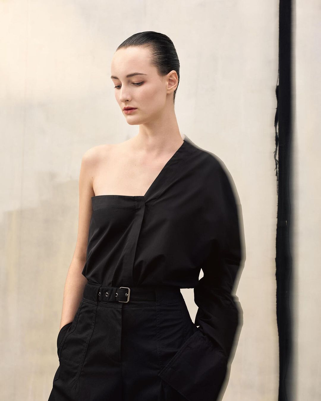 Elegant woman in a black one-shoulder top and belted trousers poses against a textured off-white background, beauty and fashion editorial.