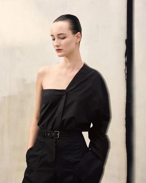 Elegant woman in a black one-shoulder top and belted trousers poses against a textured off-white background, beauty and fashion editorial.