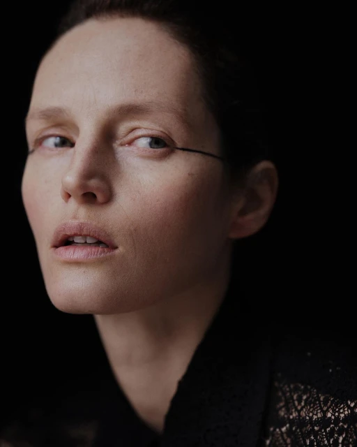 Portrait of a fair-skinned woman with minimalist makeup, dark hair, and a dark lace top against a black background, beauty shot.