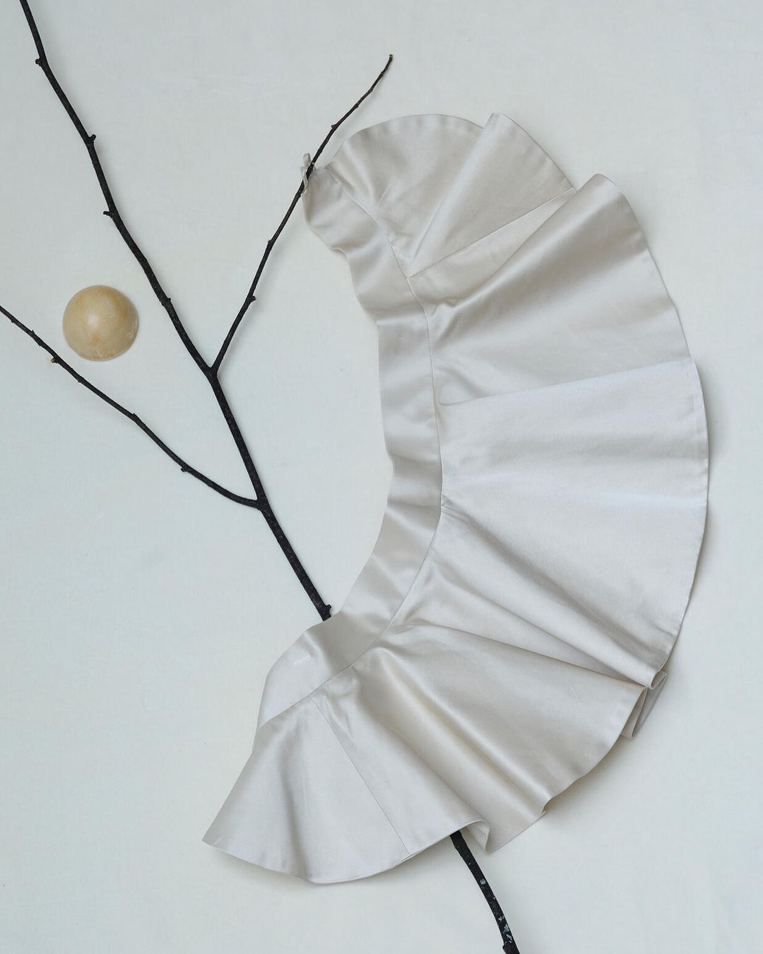 Cream-colored frilly shirt with branch and wood sphere on a white backdrop in a minimalist fashion or art advertisement.