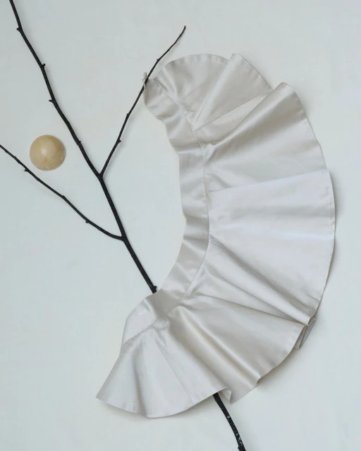 Cream-colored frilly shirt with branch and wood sphere on a white backdrop in a minimalist fashion or art advertisement.