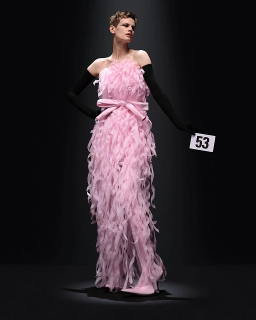 Fashion show runway featuring a model wearing a pink feather dress with a bow and black gloves under a spotlight on a black background.