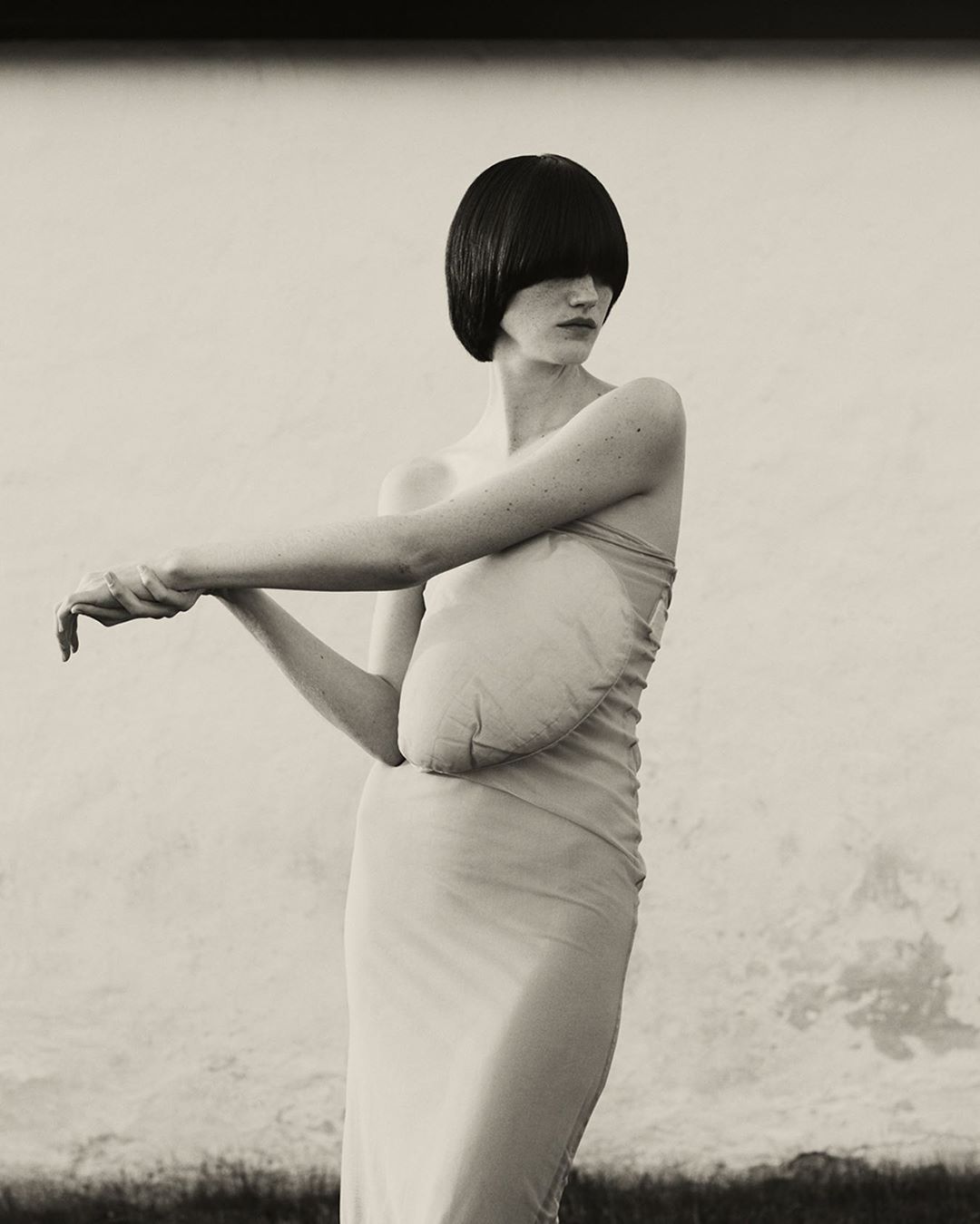 Artistic fashion shot of a woman with a dark bob haircut wearing a dress, posing against a textured wall with dark vegetation below.