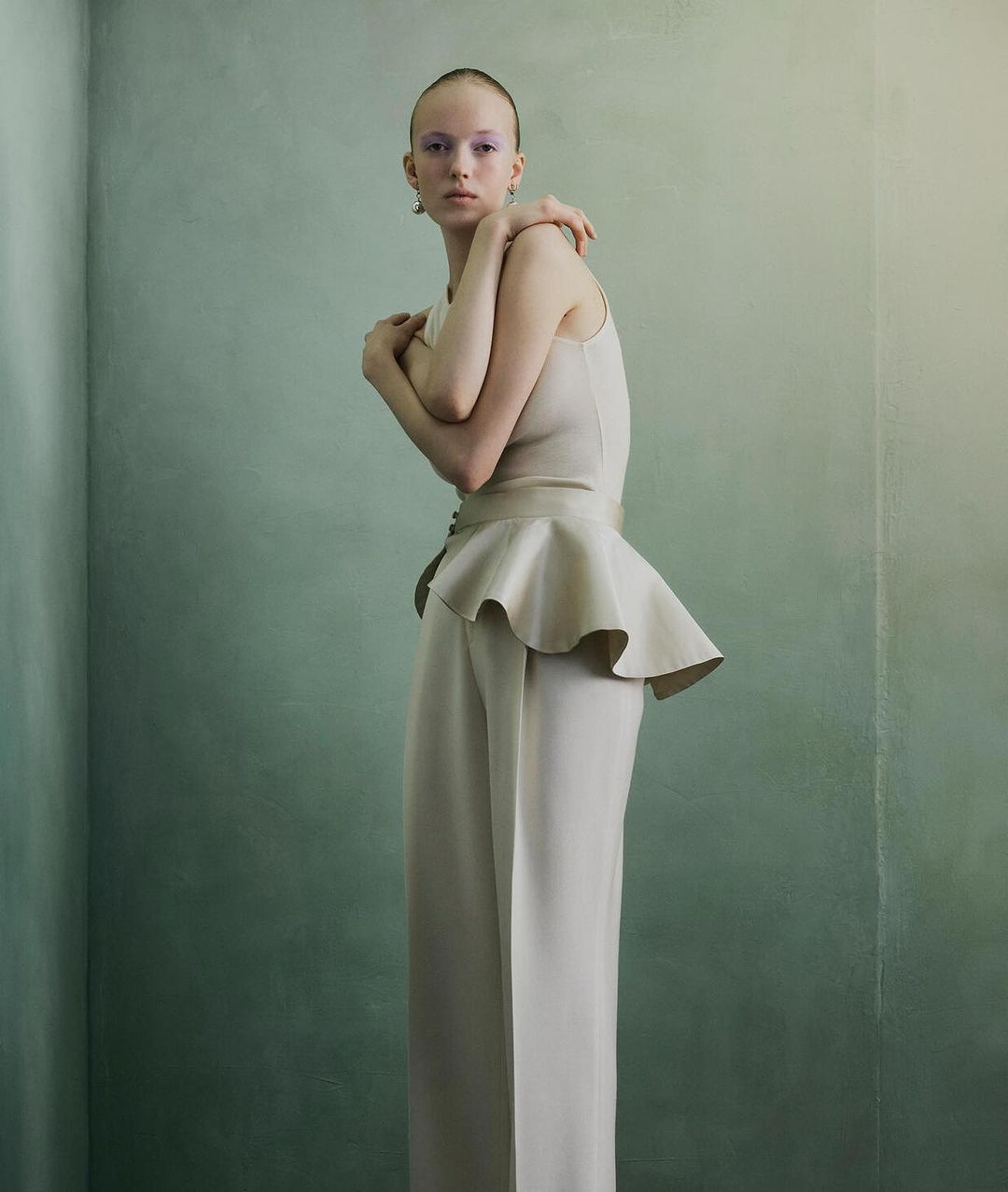 Fashion editorial shot of an adult female model wearing a light beige outfit against a painted textured wall in an indoor setting.