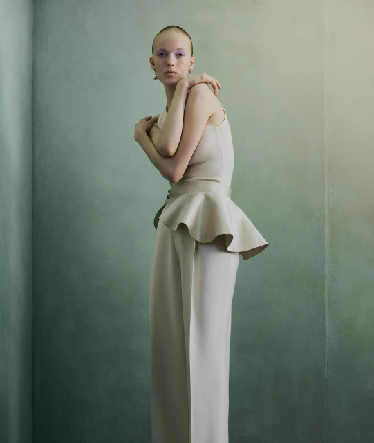 Fashion editorial shot of an adult female model wearing a light beige outfit against a painted textured wall in an indoor setting.