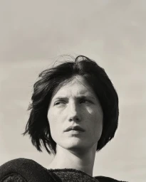 Close-up portrait of a woman with dark short hair and freckles looking off to the side against an overcast sky background