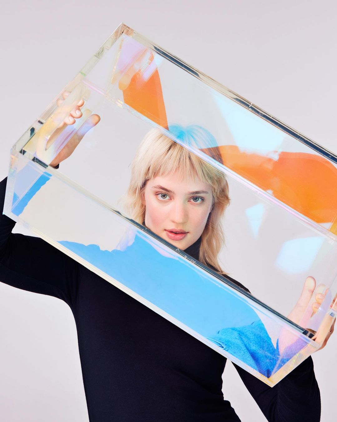 Blonde woman in black turtleneck framed by iridescent transparent rectangle, neutral background. Beauty, fashion, studio photography.