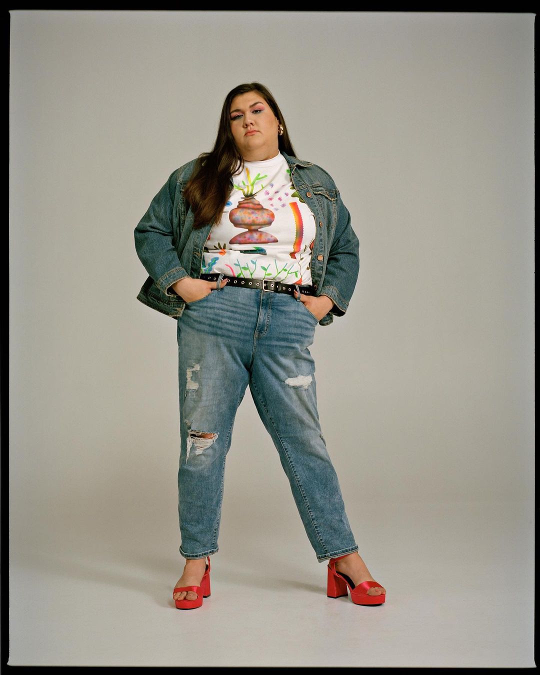 Full-length portrait of a brunette adult female in a T-shirt with a graphic design, denim jacket, ripped jeans, and platform sandals against a white background.