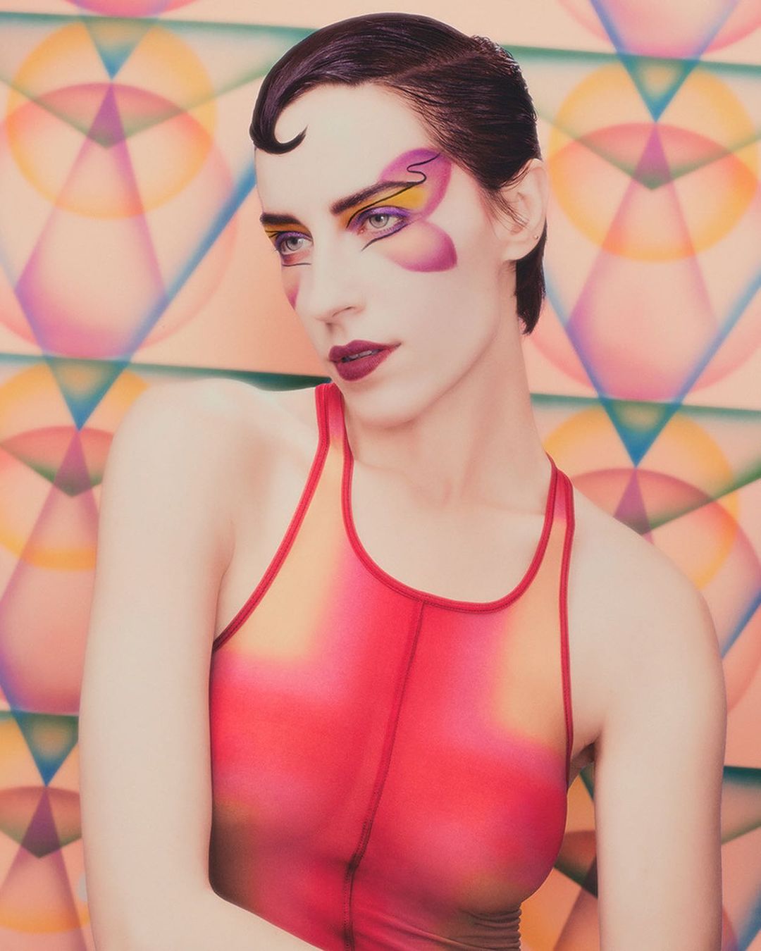 Close-up portrait of a woman with dramatic makeup and a retro hairstyle against a colorful geometric background in a studio setting.