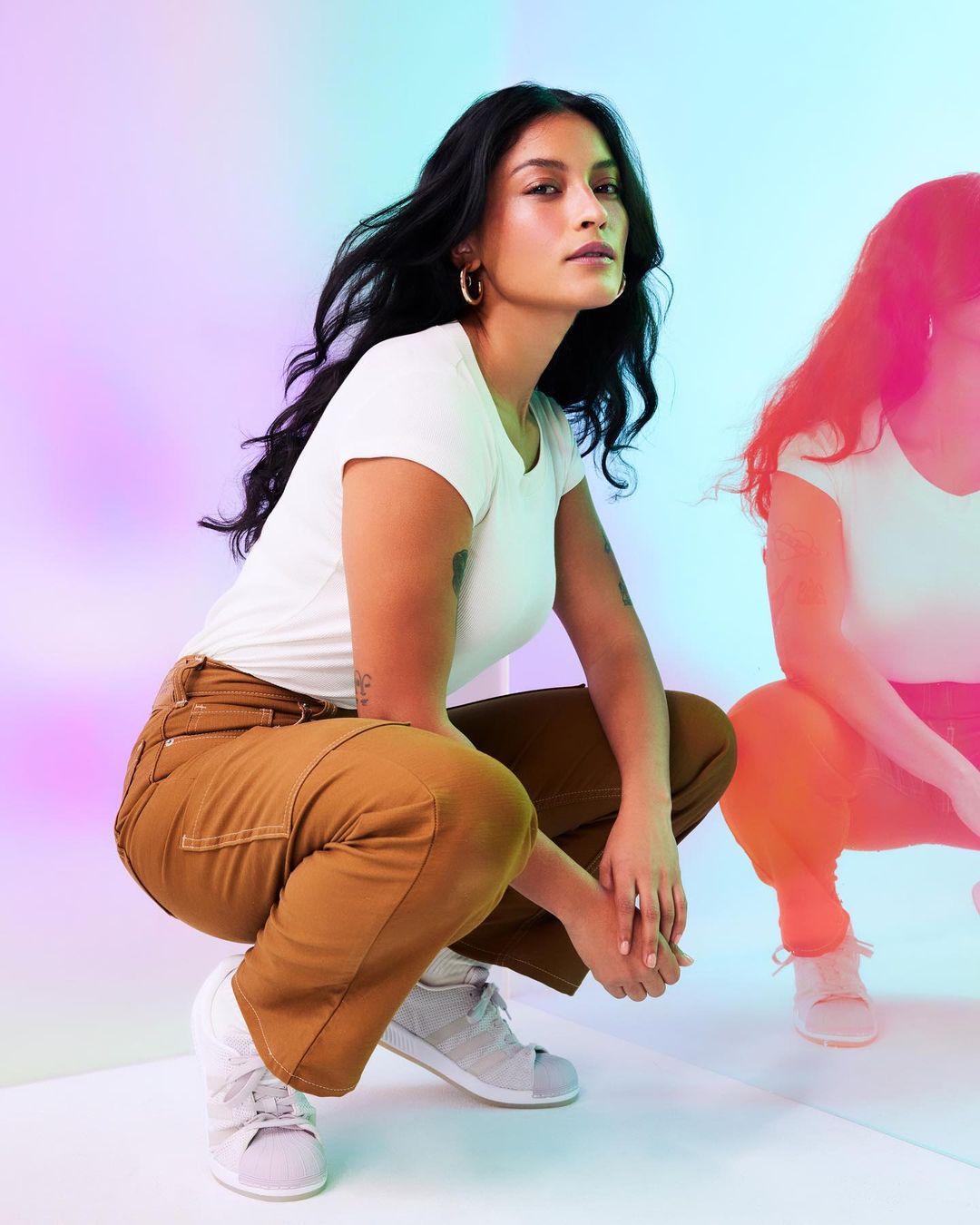 Two women pose crouching in a photo studio with a background of pastel colors. The first woman faces the camera directly.