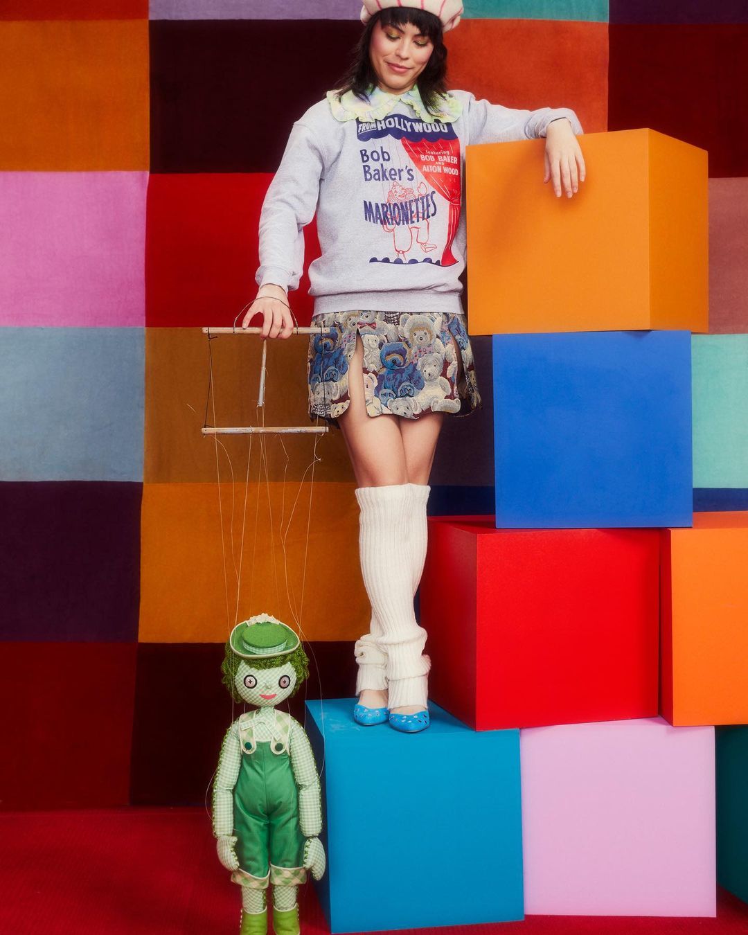 Woman poses with puppet in fashion editorial. Colorful geometric backdrop, beret, patterned skirt, knit sweater, doll-like puppet.