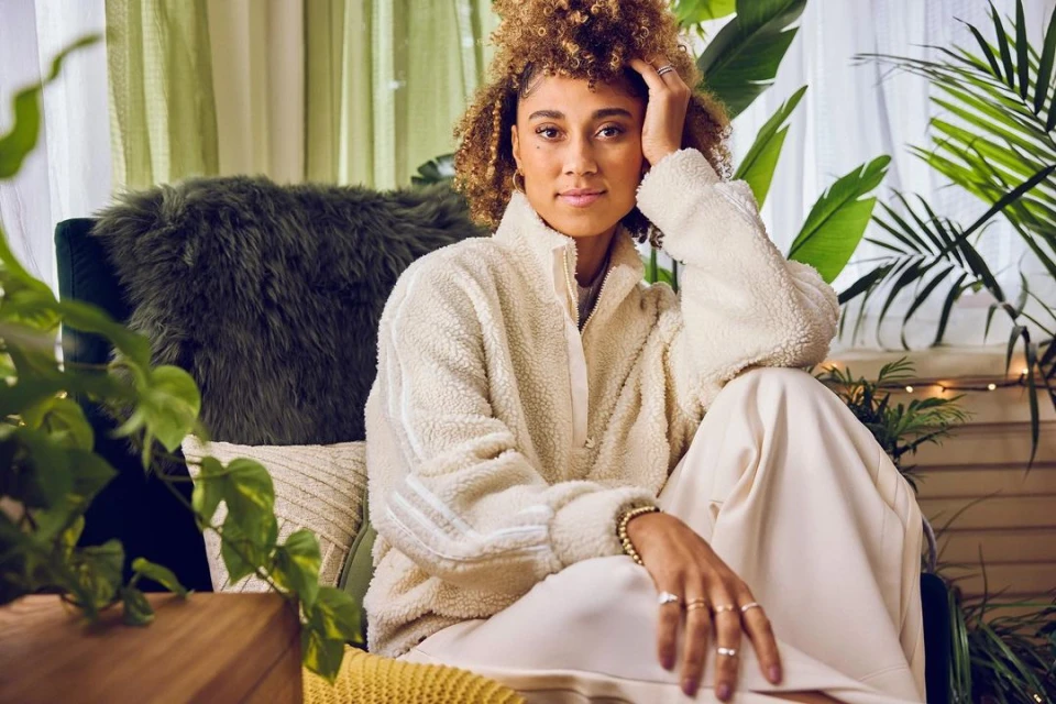 Attractive woman poses indoors with plants, wearing a white fleece jacket and cream-colored pants in a lifestyle fashion shot.