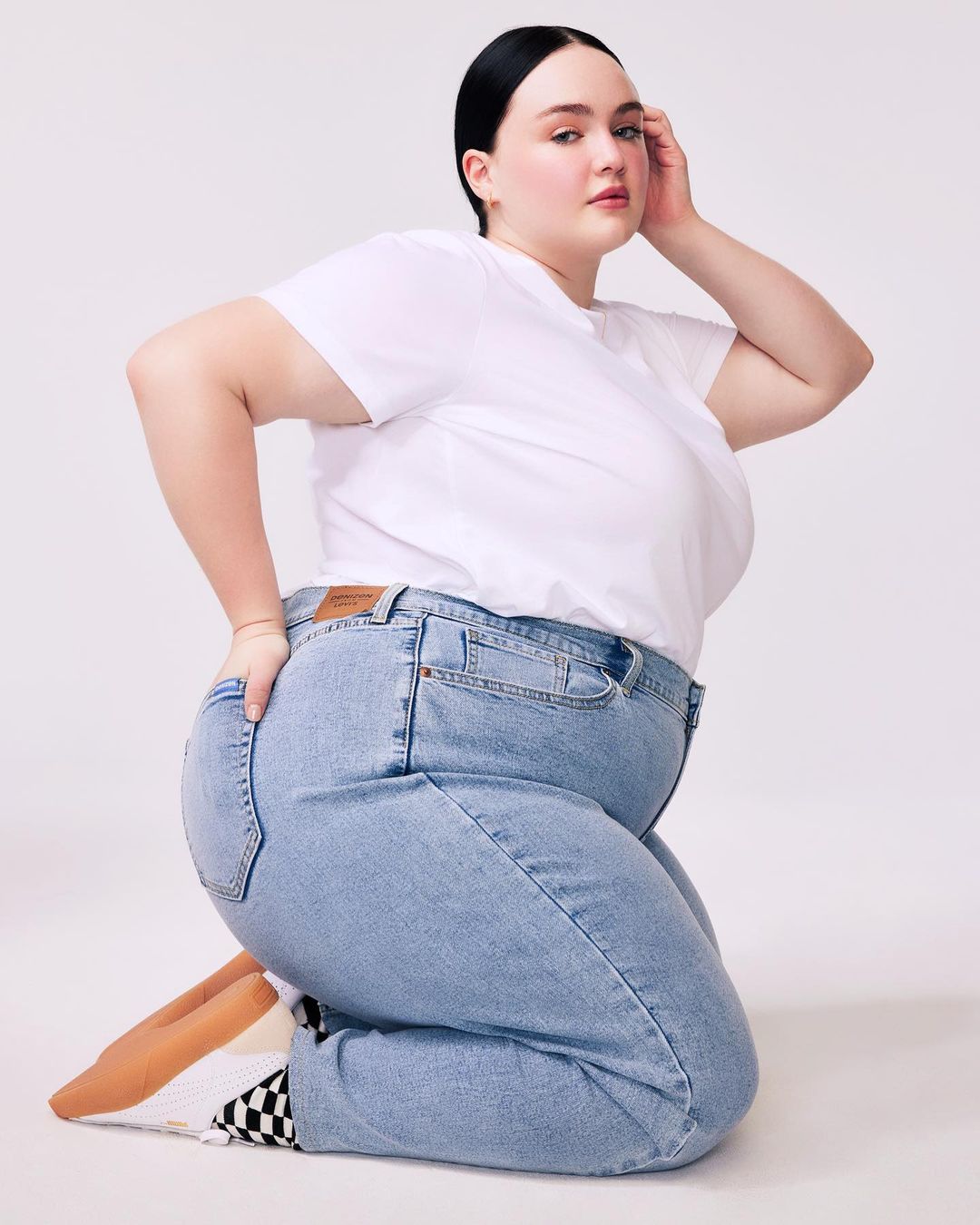 Woman in white t-shirt and jeans kneeling in a studio setting. Fashion portrait showcasing casual style, light blue denim, and simple studio backdrop.