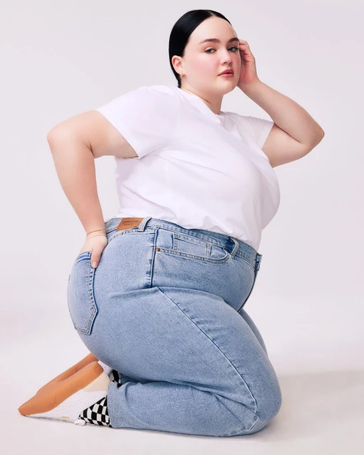 Woman in white t-shirt and jeans kneeling in a studio setting. Fashion portrait showcasing casual style, light blue denim, and simple studio backdrop.