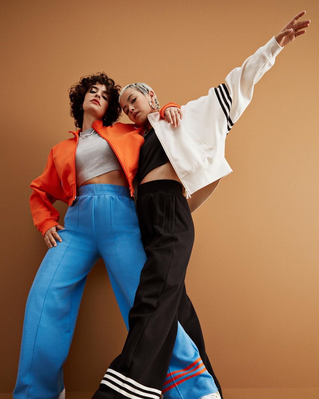 Two fashion models in Adidas-branded activewear, pose in a low angle shot against a tan backdrop, showcasing sporty jackets and pants.