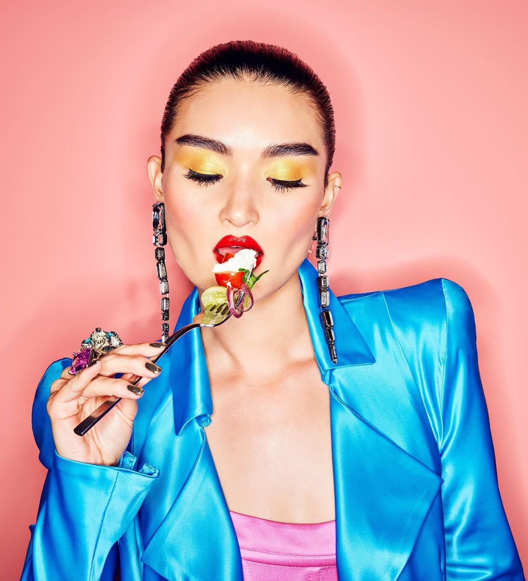 A young adult woman eats salad from a fork against a pink background. She has on striking makeup and blue jacket.