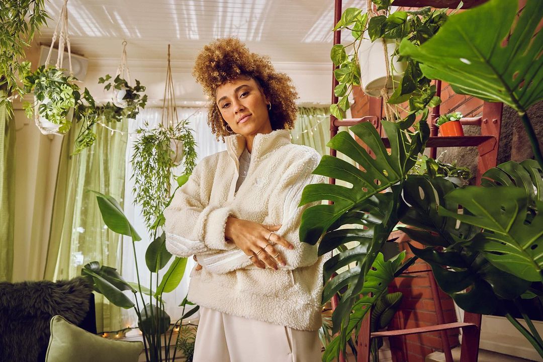 A woman with curly hair in a cream-colored jacket poses in a room filled with lush greenery, including potted and hanging plants, creating a vibrant indoor garden.