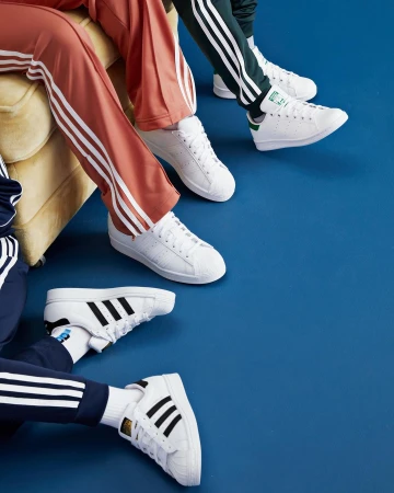 Three sets of Adidas sneakers and track pants against a blue backdrop, showing three people sitting on a yellow couch, fashion lifestyle.