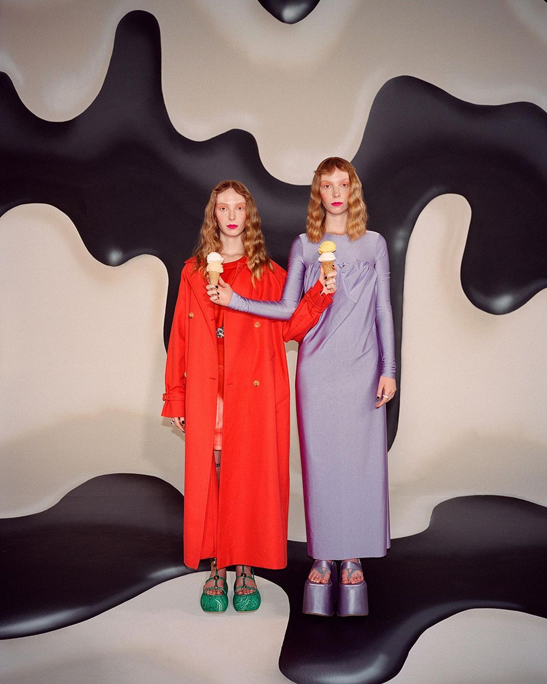 Two red-haired adult females pose in long-sleeved dresses, one red and one lilac, holding ice cream cones against an abstract backdrop.