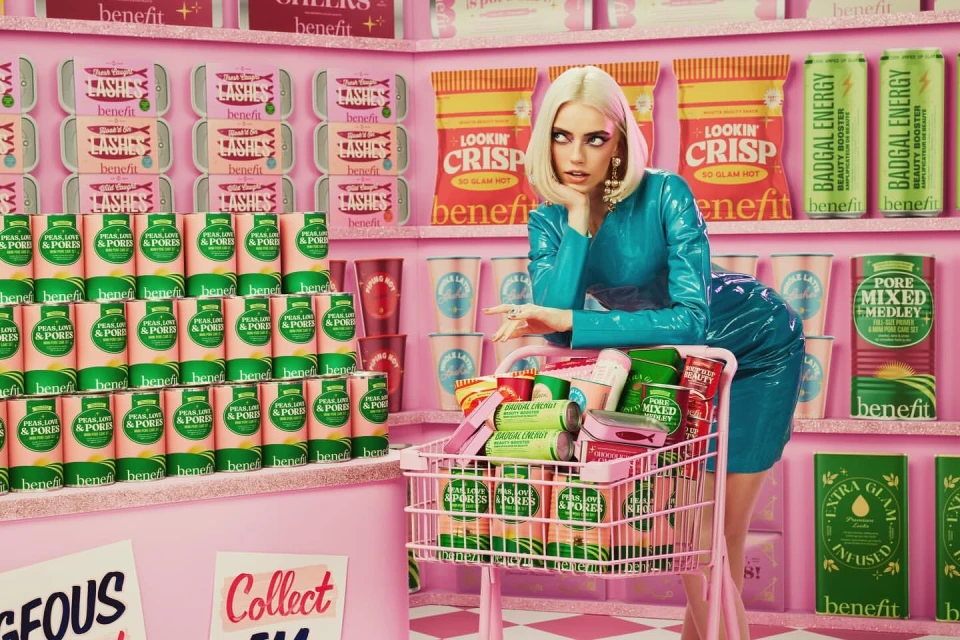 A woman poses in a pink and white beauty product grocery store, wearing a blue dress and pushing a cart full of Benefit cosmetics.