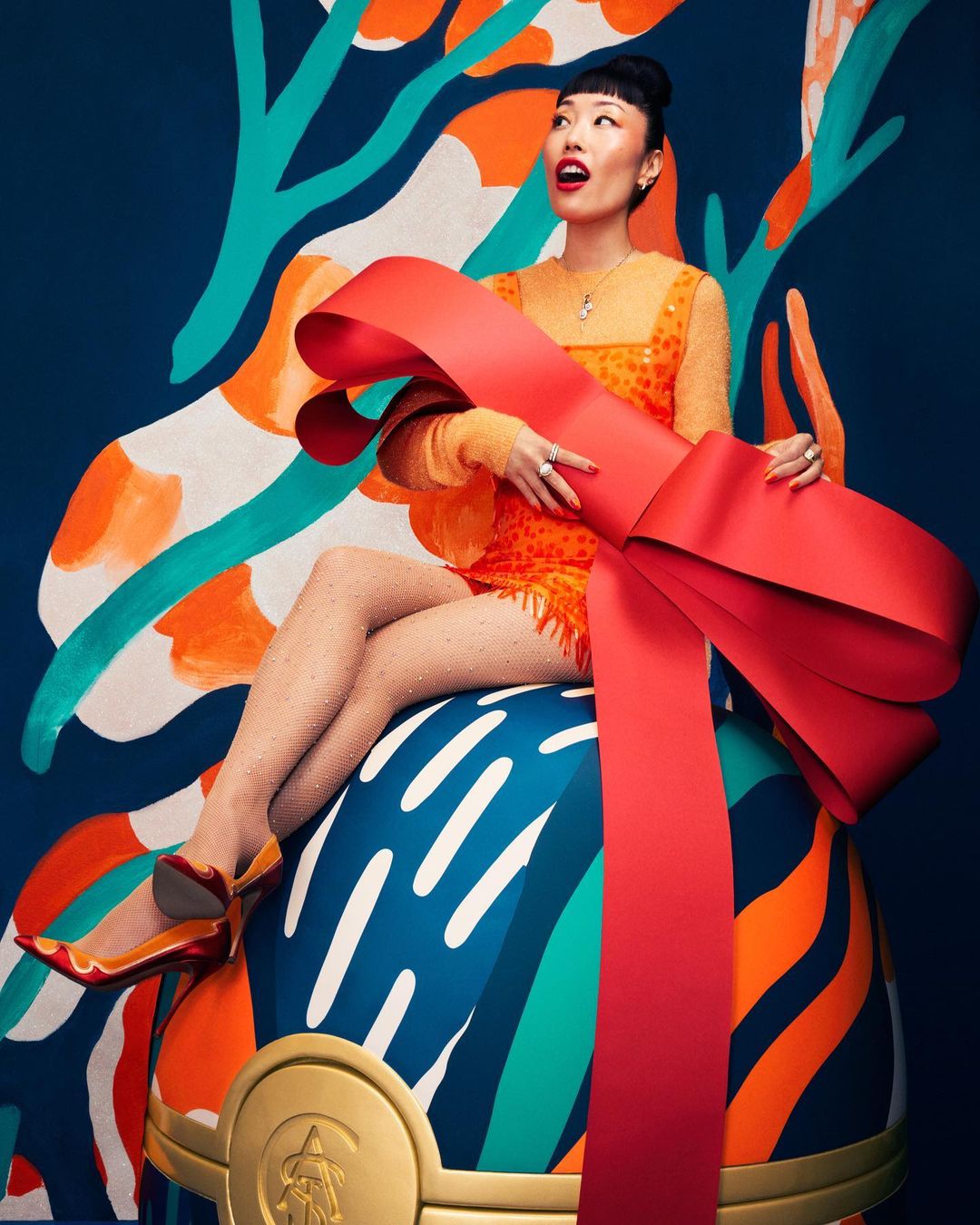 Woman sits on ornament with a bow in an orange dress and shoes with a multicolored abstract background in a studio shot.