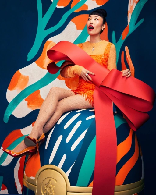 Woman sits on ornament with a bow in an orange dress and shoes with a multicolored abstract background in a studio shot.