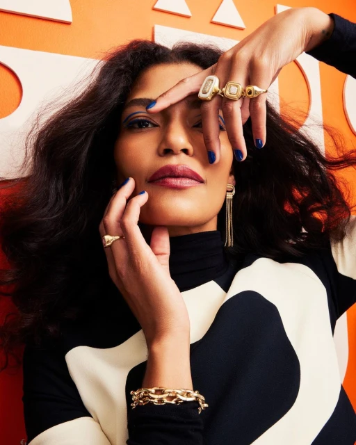 Portrait of a woman with dark hair wearing jewelry, including gold rings and a bracelet, against an orange geometric background.