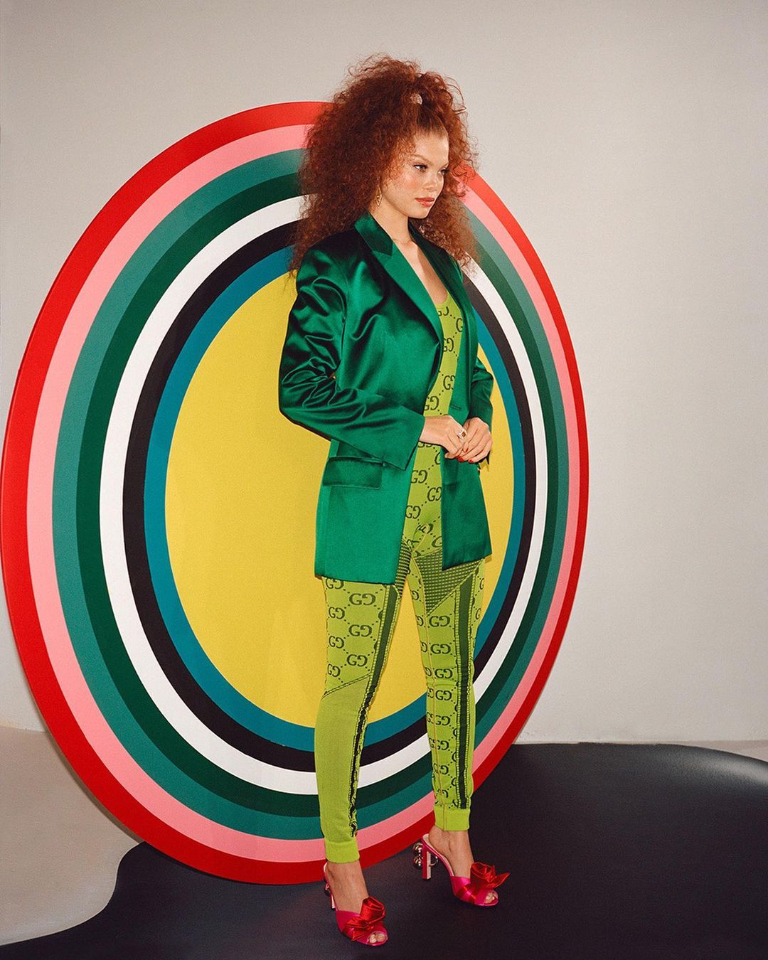 A woman with curly red hair poses in a green blazer and patterned leggings next to a colorful circular background for a fashion shoot.